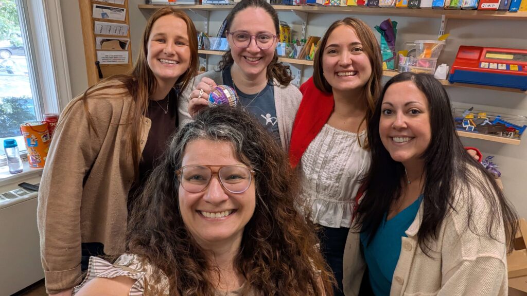 Five Kid Matters Counseling therapists smiling together in a playroom lined with games, art supplies and sensory toys.